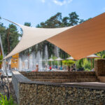 Outdoor pool area with beige fabric shade sails, stone-tiled walls, and people relaxing in the background.