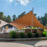 Outdoor pool area with orange and beige shade sails, people relaxing by the water fountain amid trees and greenery.