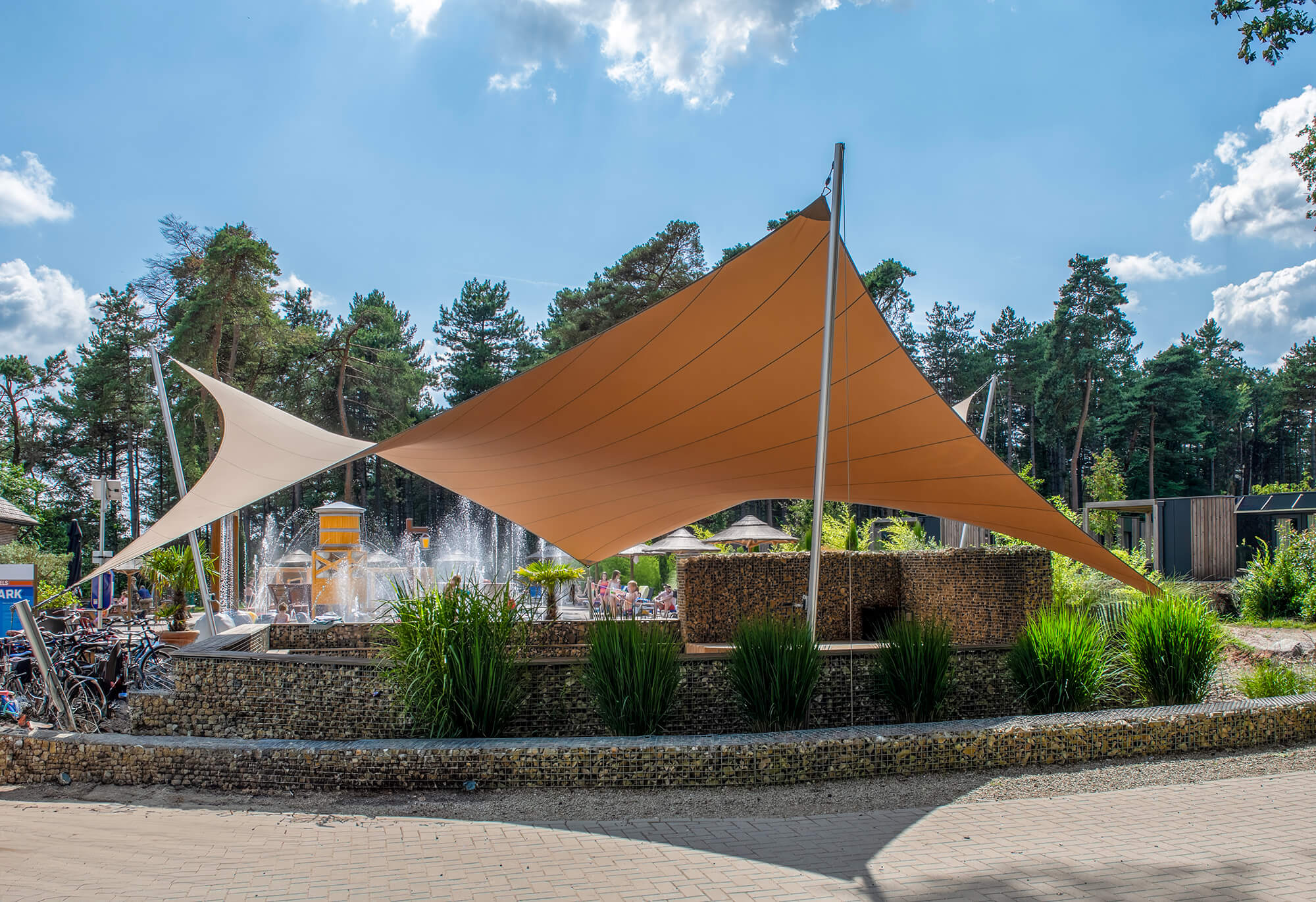 Outdoor pool area with orange and beige shade sails, people relaxing by the water fountain amid trees and greenery.