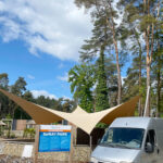 Outdoor spray park entrance with tan shade sails, trees, and a parked van nearby.