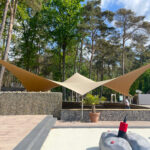 Outdoor courtyard with tan fabric shade sails stretched overhead, stone gabion walls, and a lone person walking near a building amid trees and blue sky.