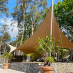 Tan triangular shade sail over a paved courtyard with potted palms and a stone retaining wall, set among tall trees under a blue sky.