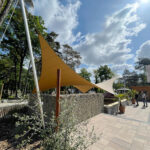 Outdoor plaza with large yellow fabric shade sails supported by metal poles, stone wall and paved walkway under a blue sky.