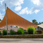 Large orange and beige fabric shade sails stretched between tall poles in front of a brick building, with greenery and bikes to the left.