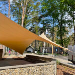 Outdoor courtyard with tan triangular shade sails, tall trees, and a gabion wall; a white van is parked to the right.