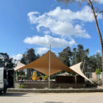 Outdoor event area with large tan fabric shade sails, stone seating wall, and surrounding trees under a blue sky with white clouds.