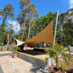 Outdoor courtyard with tan fabric shade sails, tall trees, and potted palms under a clear blue sky.