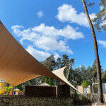 Beige fabric shade sails over an outdoor seating area with stone walls and a blue sky above tall trees.