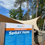 Signboard at EuroParcs Hoge Kempen showing Spray Park rules (Gedragsregels) with blue/orange panel, beige shade sail, and tall trees in the background.