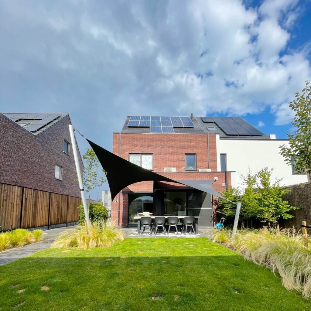 Backyard patio of a modern brick house with solar panels on the roof, black shade sails, and an outdoor dining table with chairs amid green lawn.