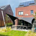 Modern backyard with red-brick house and solar panels on the roof; outdoor dining table and black chairs beneath shade sails