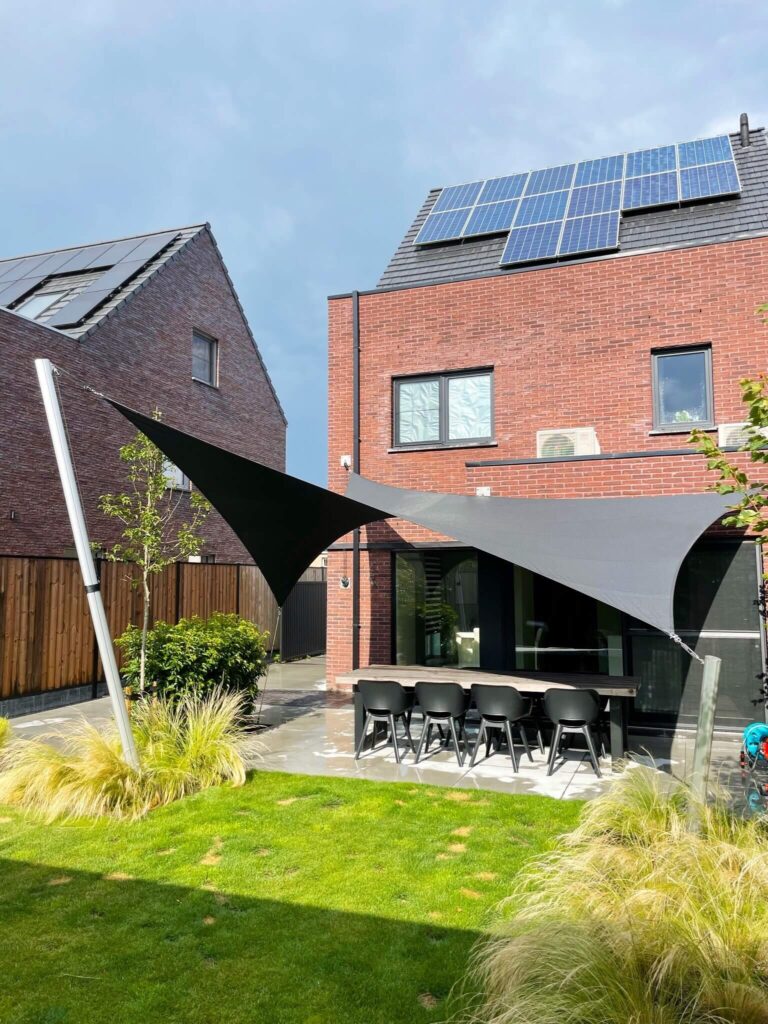 Modern backyard with red-brick house and solar panels on the roof; outdoor dining table and black chairs beneath shade sails
