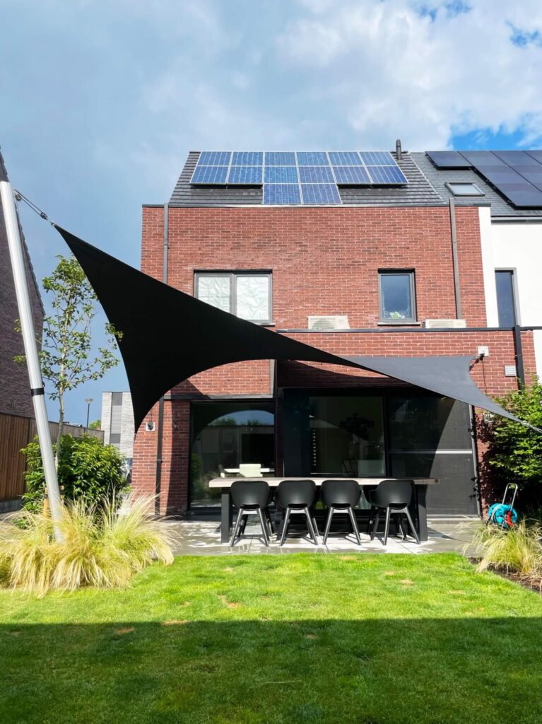 Modern brick house with solar panels on the sloped roof; outdoor dining table and chairs beneath black shade sails in the yard.