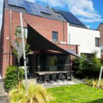 Brick house with solar panels on the roof, glass doors, and a shaded outdoor dining table in a manicured yard with plants.