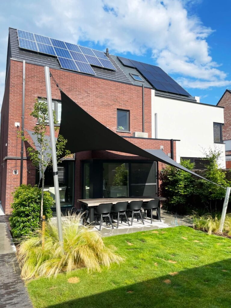 Brick house with solar panels on the roof, glass doors, and a shaded outdoor dining table in a manicured yard with plants.