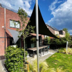 Outdoor patio area with a long wooden table and black chairs under a black shade sail beside a red brick house with solar panels on the roof.