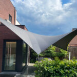 Gray triangular shade sail stretched between brick house and posts over a patio area with glass door and lush greenery, bright blue sky above