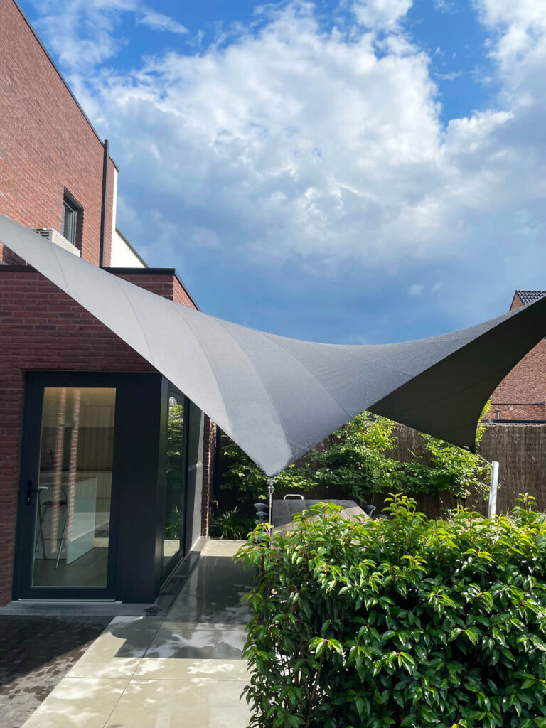 Gray triangular shade sail stretched between brick house and posts over a patio area with glass door and lush greenery, bright blue sky above