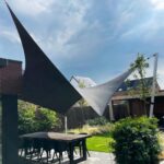 Outdoor patio with black triangular shade sails anchored to a structure and a tall pole, table and chairs beneath, garden in background
