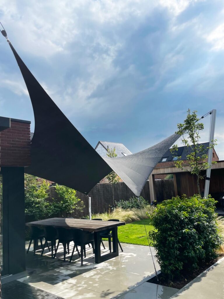 Outdoor patio with black triangular shade sails anchored to a structure and a tall pole, table and chairs beneath, garden in background
