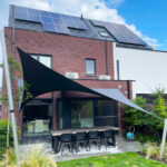 Backyard patio of a modern brick house with solar panels on the roof; black shade sails over a long dining table and eight chairs.