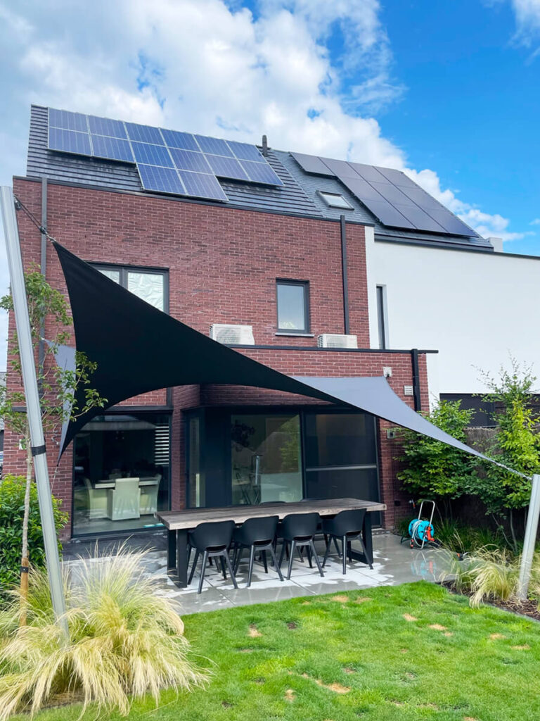 Backyard patio of a modern brick house with solar panels on the roof; black shade sails over a long dining table and eight chairs.