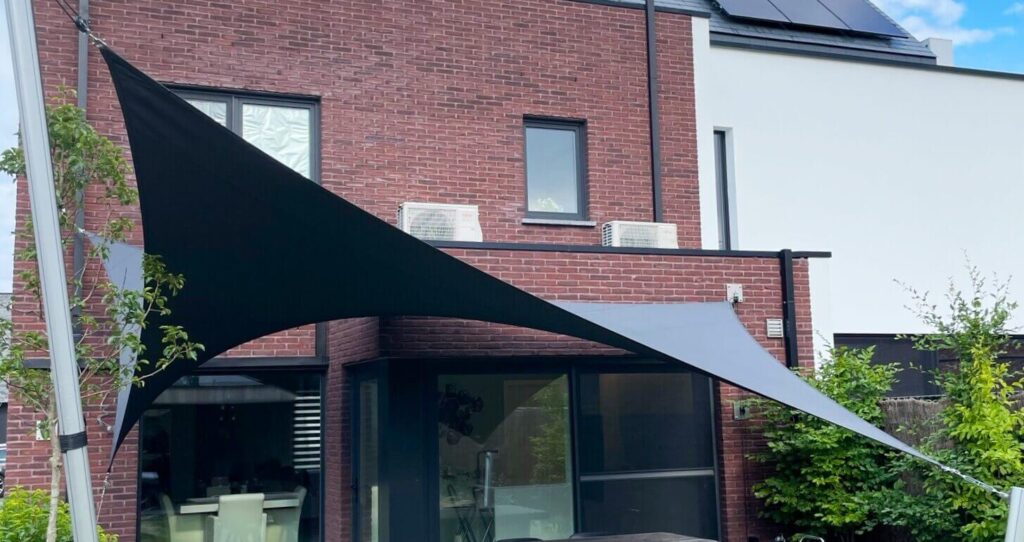 Backyard patio with black and gray fabric shade sails stretched between posts above a glass door and dining area in a brick house.