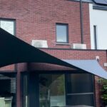Backyard patio with black and gray fabric shade sails stretched between posts above a glass door and dining area in a brick house.