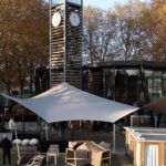 Outdoor seating area under white fabric shade sails in front of a glass-walled modern building, with autumn trees in the background and people standing around.