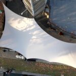 Overhead view of a large curved white fabric canopy spanning a paved courtyard with fallen autumn leaves.