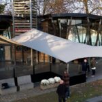 Contemporary glass-and-wood building with a large white tensile shade sail and a small group of people outside on a fall day near a body of water.