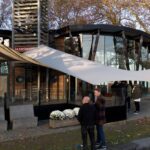 Modern glass pavilion with a large white sail shade, people gathered outside on a fall day by a river.