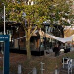 Riverside glass pavilion with a white sail canopy, people chatting near the dock at Port de plaisance de Visé amid autumn trees.