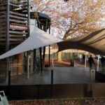 Outdoor cafe terrace beside a lake with white and brown tension shade sails, a wooden building labeled 'La Capitainerie', and autumn trees in the background.