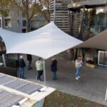 Group of people standing and chatting under a large white tension fabric shade sail outside a glass-front building, with solar panels in the foreground.