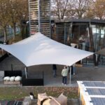 Large white tensile canopy over a courtyard patio in front of a glass-walled building, with people underneath.