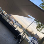 Harbor scene with moored boats along the water, viewed through a glass railing under a large tan fabric shade sail.