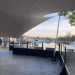 Outdoor marina area with a large curved fabric shade canopy over seating and a calm waterway with boats in the distance.