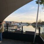 Outdoor riverside seating area under a large curved canopy with boats docked along a calm river.