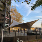 Outdoor seating area with a large two-tone tensile shade sail, glass railing, and a wooden lattice tower on the left; autumn trees and people nearby.