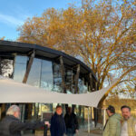 Group of adults standing outside a glass-front building under a white curved canopy, with autumn trees and blue sky visible.