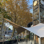 Outdoor courtyard with autumn-colored trees, beige shade sails, and a tall wooden lattice tower with shop signs against a blue sky.
