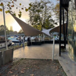 Marina promenade with moored boats, people strolling under tan and white shade sails at sunset.
