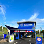 Blue carwash building with a large 'Carwash' sign and a car entering the wash bay under a red-and-white barrier on a sunny day.