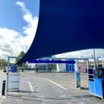 Blue curved shade sail covering a service plaza with blue kiosks and white buildings in the background, outdoor pavement visible.