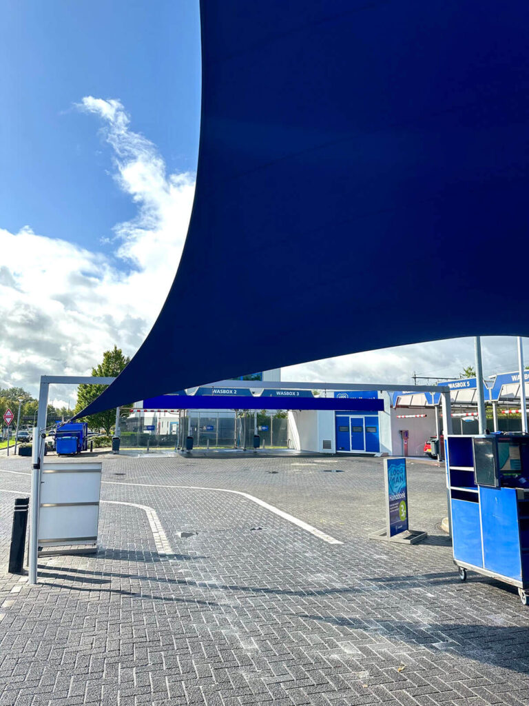 Blue curved shade sail covering a service plaza with blue kiosks and white buildings in the background, outdoor pavement visible.