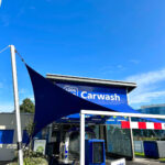 Blue car wash entrance under a large blue shade sail, with a payment kiosk and a prominent 'Carwash' sign.