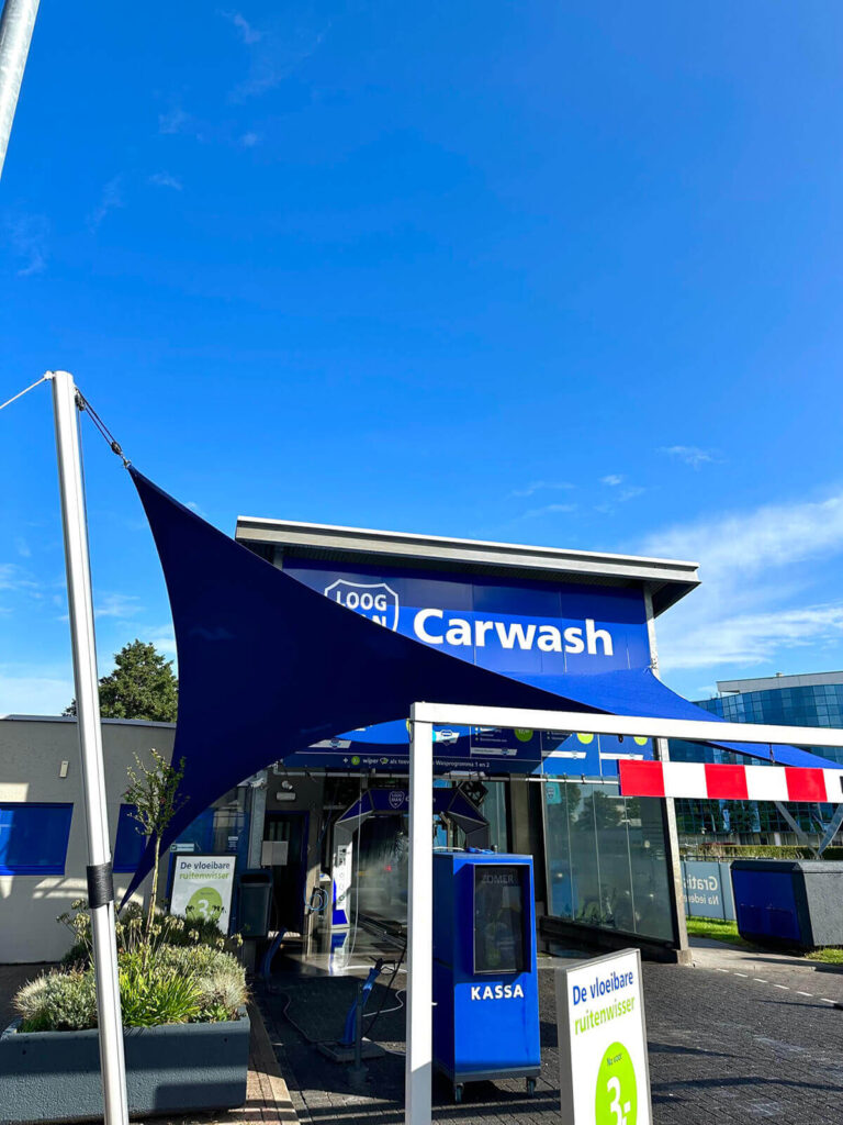 Blue car wash entrance under a large blue shade sail, with a payment kiosk and a prominent 'Carwash' sign.