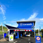 Blue and white Log Man Carwash building with a car entering the wash bay under a blue canopy; prominent 'Carwash' sign nearby.