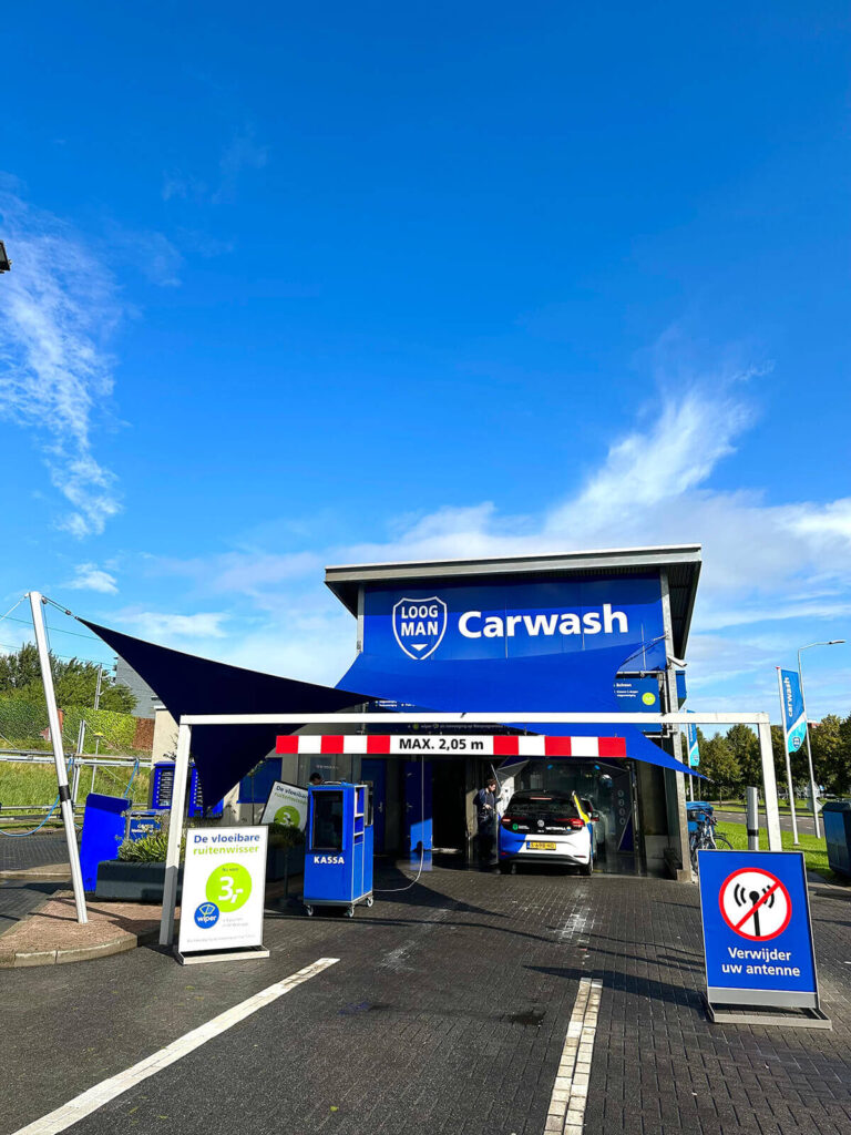Blue and white Log Man Carwash building with a car entering the wash bay under a blue canopy; prominent 'Carwash' sign nearby.